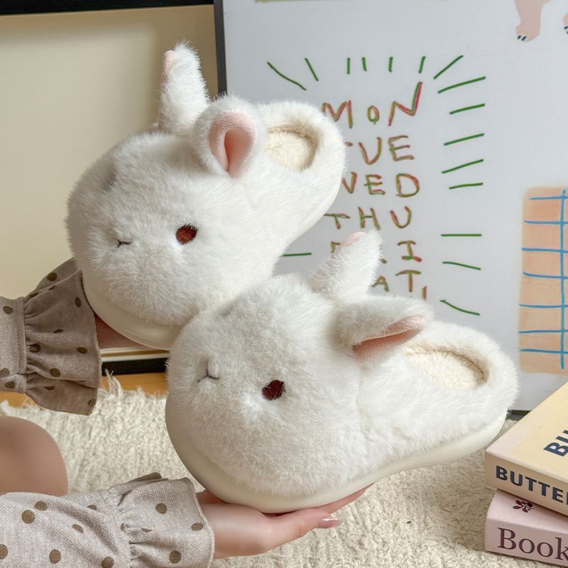Pair of white bunny plush slippers with pink lined ears held in two hands in front of a printed poster