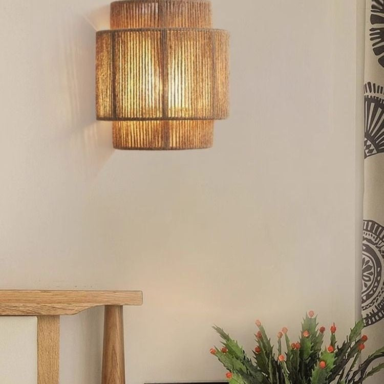 Lit stacked-tier hemp rope wall lamp on a pale wall beside a curtain, a wooden stool and a green plant
