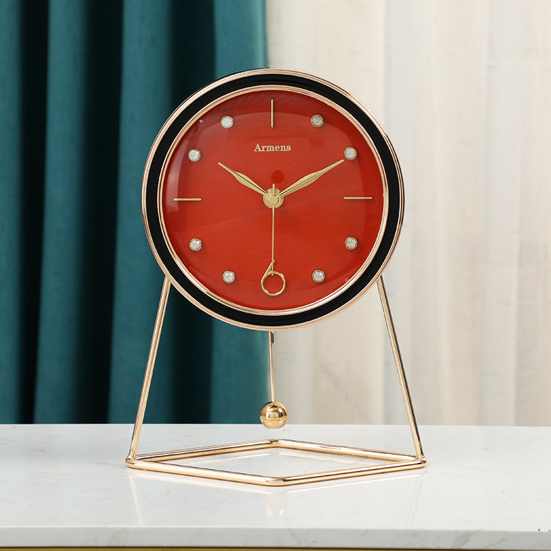 Orange dial pendulum desk clock with black bezel, gold hands and a gold ball pendulum on a white table