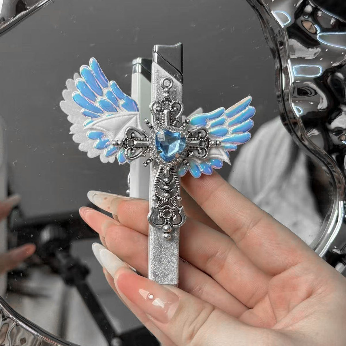 Hand holding the angel wings cross lighter upright before a mirror, white wings and blue heart stone visible
