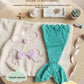 Flat lay of the three piece mermaid set, teal tail with pink tie back top and flower headband on a blanket