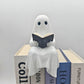 White resin ghost reading a dark book, perched on a stack of hardcover books against a pale wall