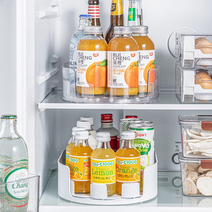 Clear oval rotating tray of orange juice bottles on a refrigerator shelf above a white tray holding vitamin drinks