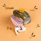 Olive frog case with a gold crown open beside a pink lower shell and a pair of AirPods Pro