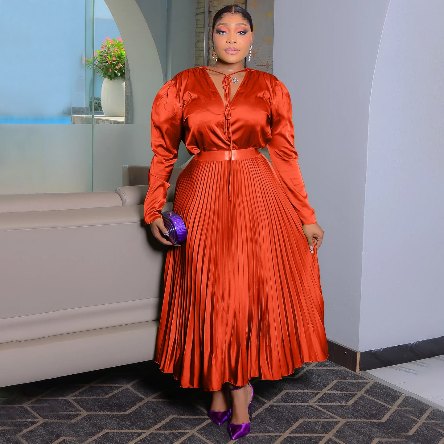 Burnt orange high shine pleated maxi dress with tie neck and matching belt, worn indoors