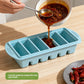 A spoon pouring red chili broth from a bowl into a light blue six compartment silicone tray on a wooden table