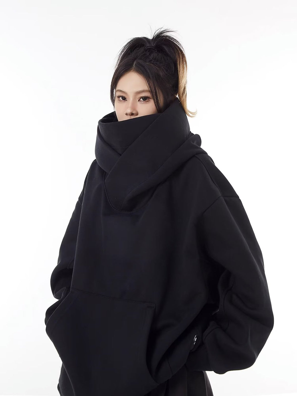 Black oversized hooded sweatshirt with the tall stand collar pulled up over the chin and a hand in the front pocket