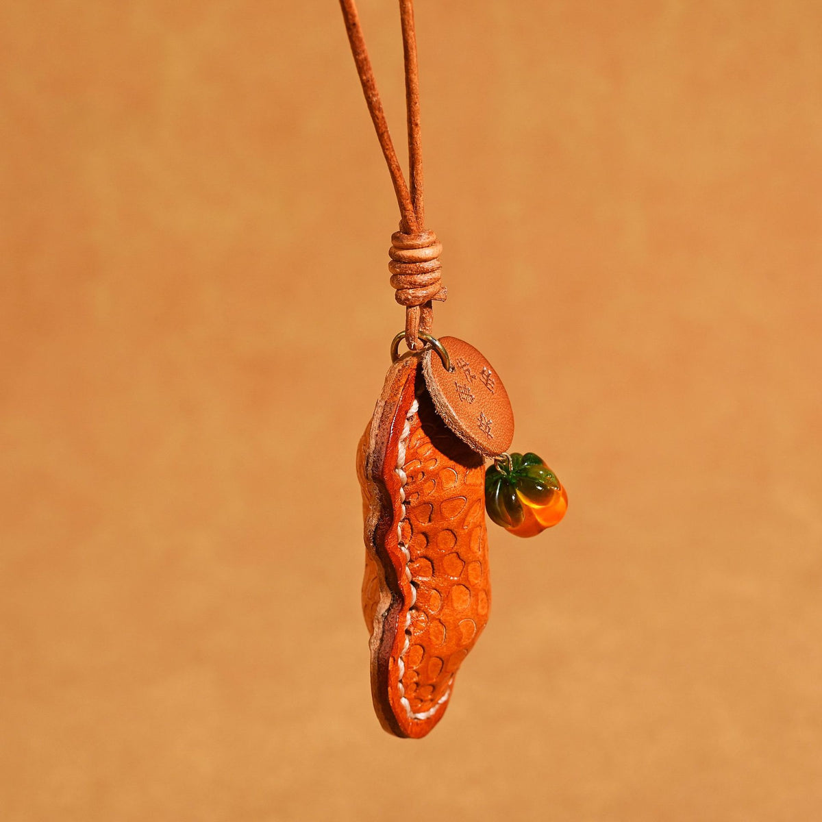 Tan orange peanut shaped leather pendant hanging from a knotted cord with an orange glass bead and round stamped tag