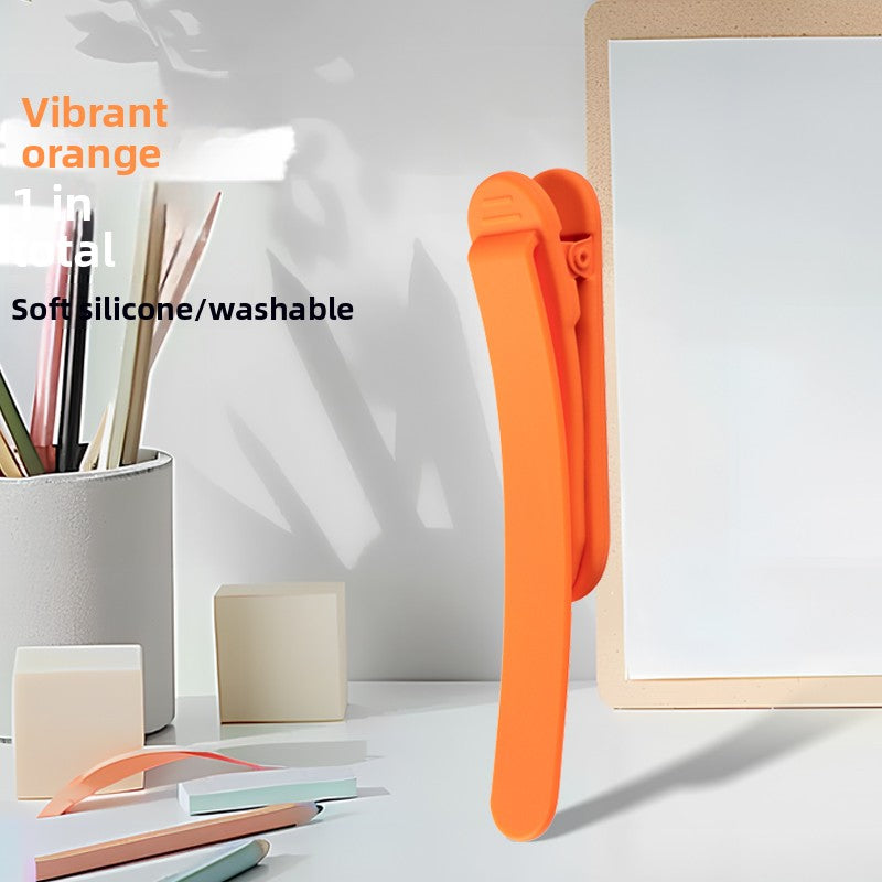 Single vibrant orange silicone bookmark clip standing on a desk beside a pen cup