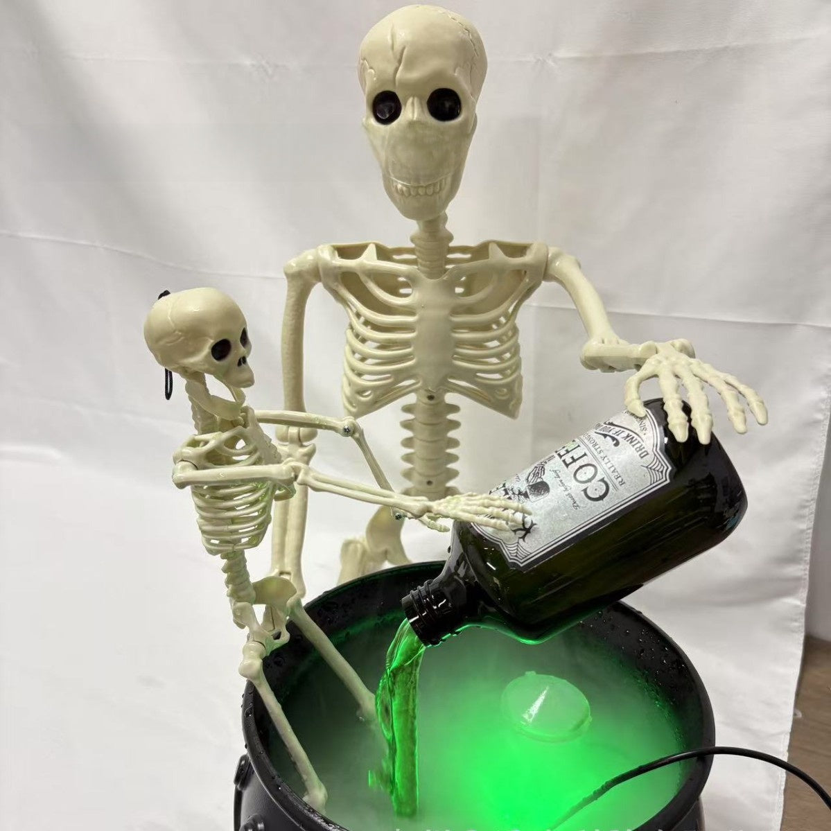 Large and small skeleton figures around a black cauldron with a tilted bottle pouring green liquid and mist
