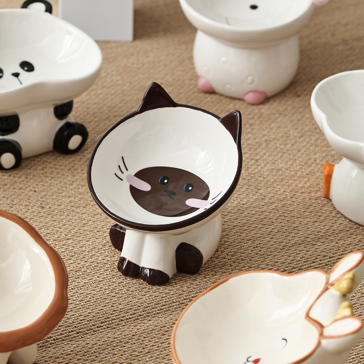 Siamese cat shaped raised ceramic bowl on a woven mat next to panda and other ceramic bowls