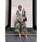 Woman looking down in green camouflage wide-leg pants with a striped top and a black bag, standing on a paved street