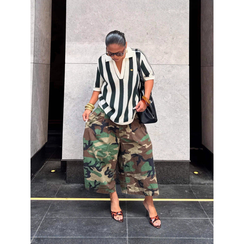 Woman looking down in green camouflage wide-leg pants with a striped top and a black bag, standing on a paved street