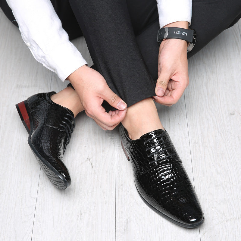 Man seated on a pale floor tying the laces of a black croc embossed derby shoe, dark trousers and white shirt