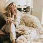 Woman in a wide-brim hat sitting on a sofa with a phone and a ruched cream plush blanket spread beside her