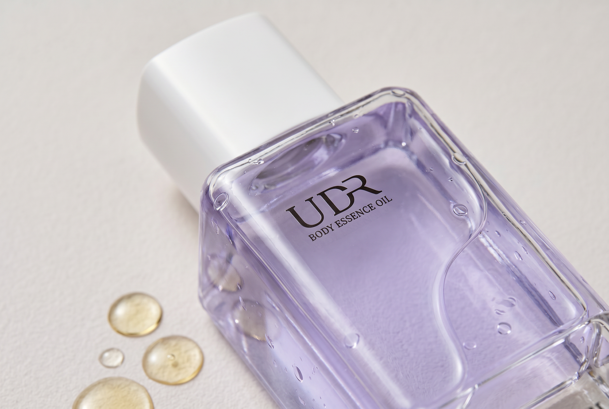 Close up of the UDR body essence oil bottle showing golden oil droplets beside it