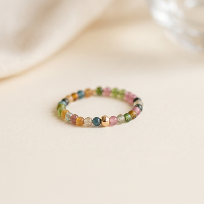 Delicate rainbow tourmaline beaded stretch ring with one gold accent bead on a cream surface