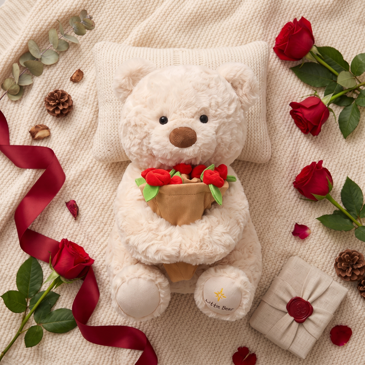 Flatlay of the cream plush rose bear with real red roses, ribbon and a wrapped gift